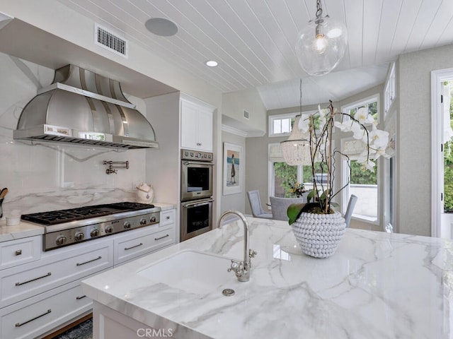 kitchen featuring stainless steel appliances, wall chimney range hood, light stone counters, pendant lighting, and white cabinets