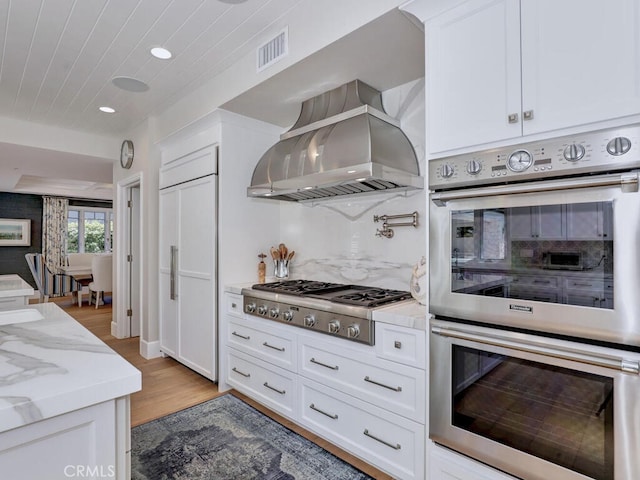 kitchen featuring light stone countertops, appliances with stainless steel finishes, wall chimney exhaust hood, light hardwood / wood-style floors, and white cabinetry