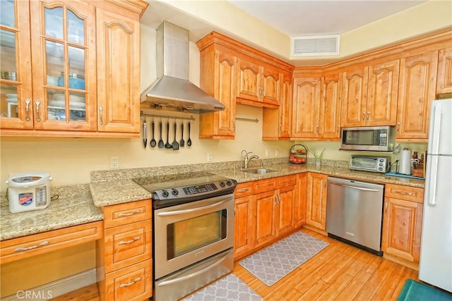 kitchen with appliances with stainless steel finishes, light wood-type flooring, light stone countertops, wall chimney exhaust hood, and sink