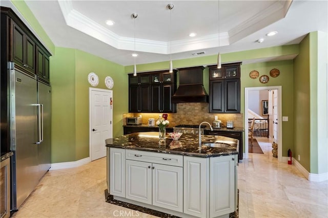 kitchen featuring tasteful backsplash, built in refrigerator, an island with sink, a tray ceiling, and custom exhaust hood