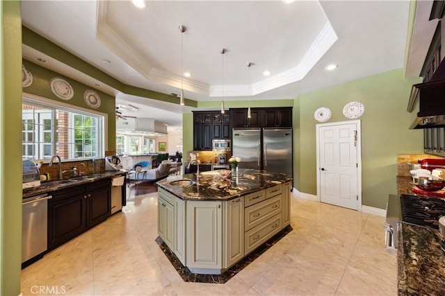 kitchen featuring ceiling fan, a raised ceiling, dark stone counters, a kitchen island with sink, and appliances with stainless steel finishes