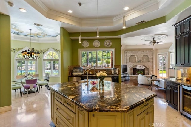 kitchen featuring a raised ceiling, dark stone countertops, an island with sink, a fireplace, and ceiling fan with notable chandelier