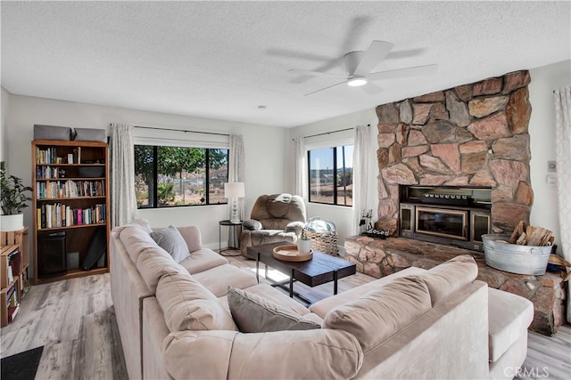 living room with light wood-type flooring, a stone fireplace, ceiling fan, and a textured ceiling