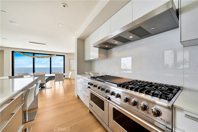 kitchen with wall chimney range hood, light wood-type flooring, double oven range, white cabinetry, and a water view