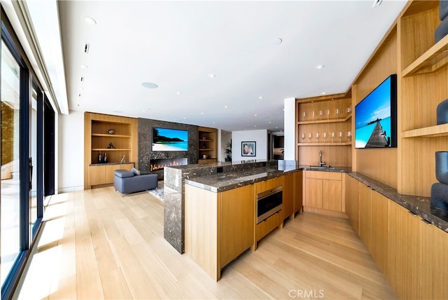 kitchen with a large fireplace, sink, dark stone countertops, built in features, and light wood-type flooring