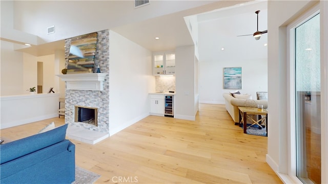 living room with a fireplace, beverage cooler, bar, ceiling fan, and light hardwood / wood-style flooring