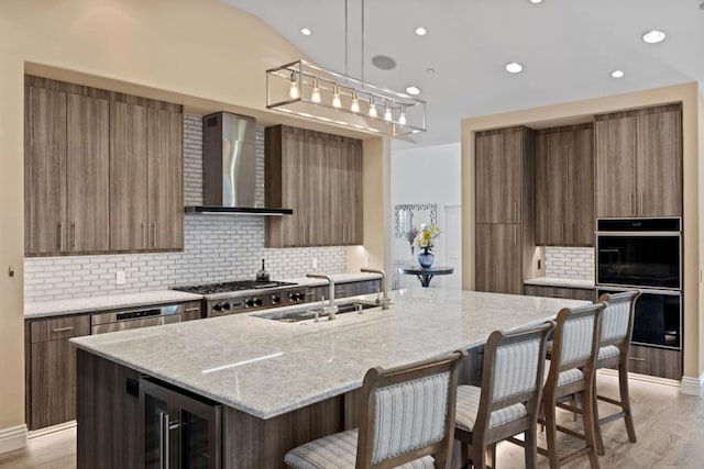 kitchen featuring a center island with sink, wall chimney range hood, black double oven, wine cooler, and decorative light fixtures