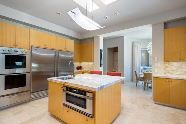 kitchen featuring stainless steel appliances, a center island with sink, tasteful backsplash, light stone counters, and a skylight