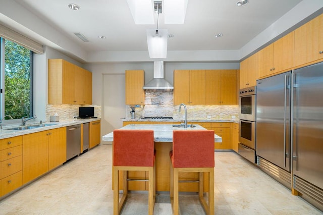 kitchen featuring a skylight, sink, stainless steel appliances, wall chimney exhaust hood, and a center island with sink