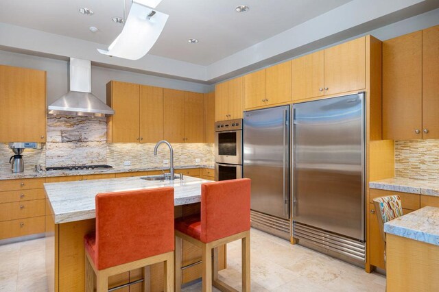 kitchen featuring wall chimney range hood, appliances with stainless steel finishes, sink, an island with sink, and decorative backsplash