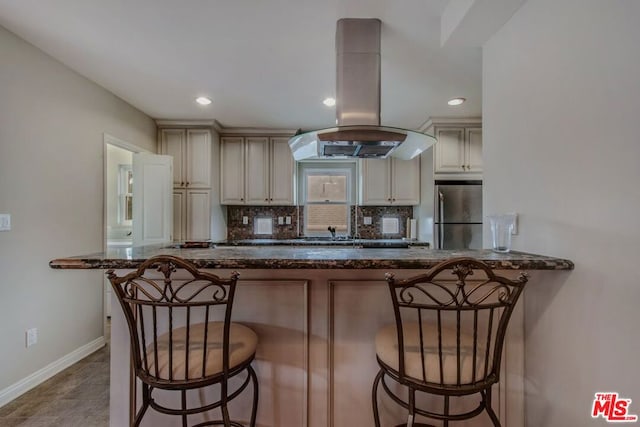 kitchen featuring dark stone counters, stainless steel fridge, cream cabinetry, a kitchen bar, and island exhaust hood