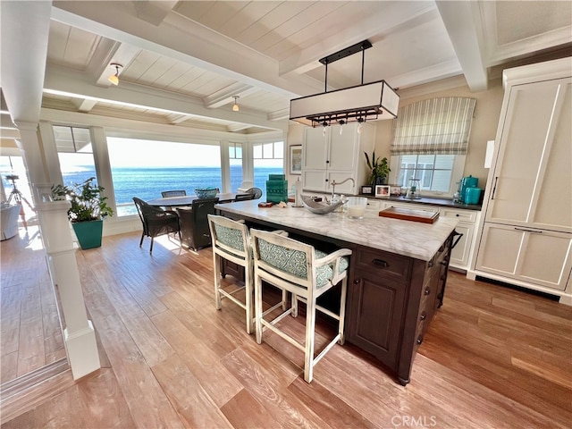 kitchen with beam ceiling, a water view, dark brown cabinetry, and an island with sink