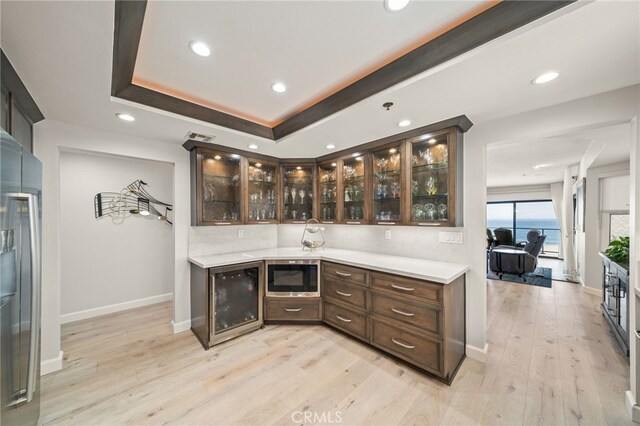 bar featuring stainless steel microwave, wine cooler, a tray ceiling, dark brown cabinets, and light wood-type flooring