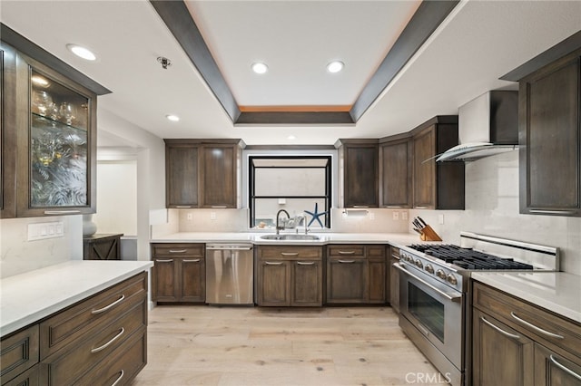 kitchen with a raised ceiling, stainless steel appliances, light countertops, wall chimney range hood, and a sink