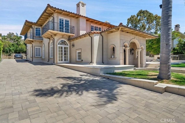 back of property with french doors, a chimney, stucco siding, a balcony, and decorative driveway