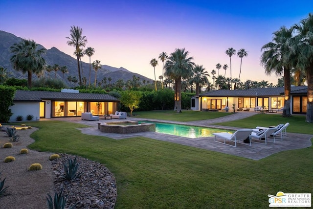 pool at dusk with a patio, a mountain view, an in ground hot tub, and a lawn