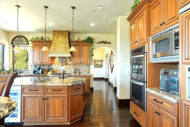 kitchen with light stone countertops, premium range hood, stainless steel appliances, arched walkways, and decorative backsplash