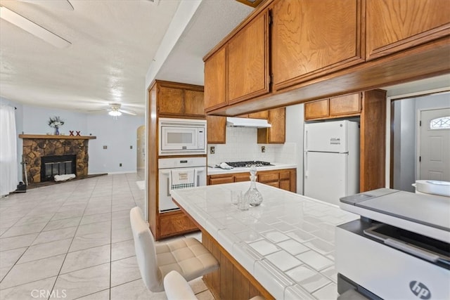 kitchen featuring decorative backsplash, a stone fireplace, tile counters, white appliances, and ceiling fan