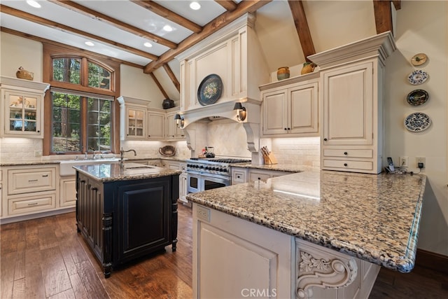 kitchen with range with two ovens, light stone countertops, beamed ceiling, and an island with sink