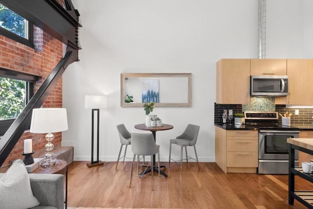 kitchen with light brown cabinets, a towering ceiling, light hardwood / wood-style floors, decorative backsplash, and appliances with stainless steel finishes