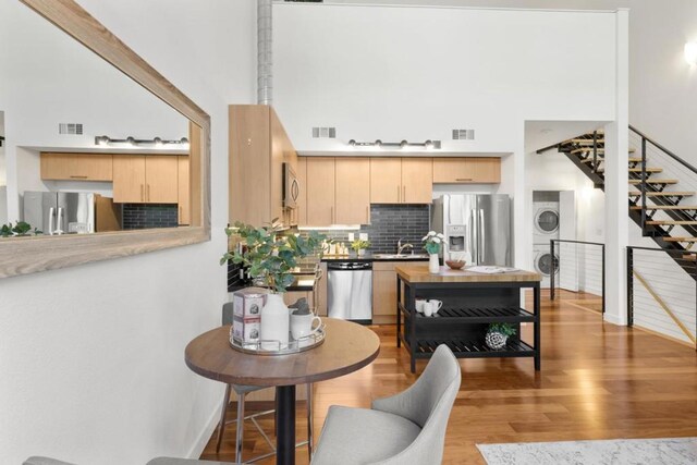 kitchen with light brown cabinetry, a towering ceiling, light hardwood / wood-style floors, stainless steel appliances, and stacked washer and dryer