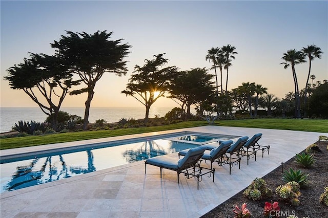 pool at dusk with a lawn, a patio area, a water view, and an in ground hot tub