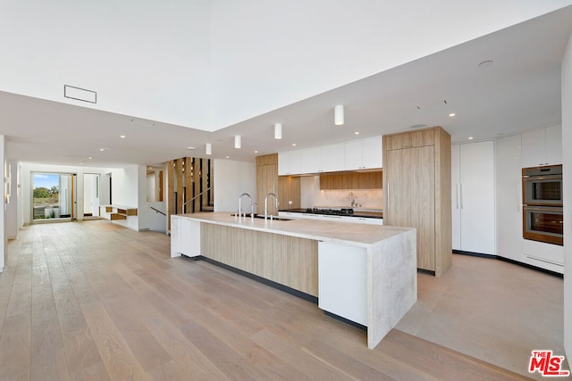 kitchen with white cabinetry, stainless steel double oven, tasteful backsplash, an island with sink, and light hardwood / wood-style floors