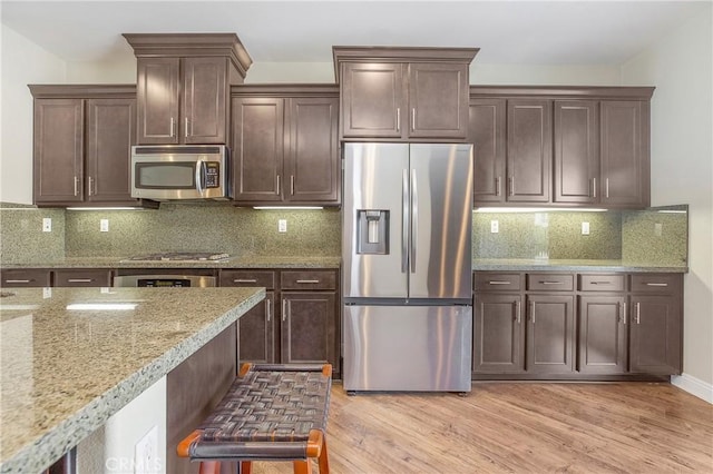 kitchen featuring tasteful backsplash, light wood-type flooring, light stone countertops, stainless steel appliances, and dark brown cabinets