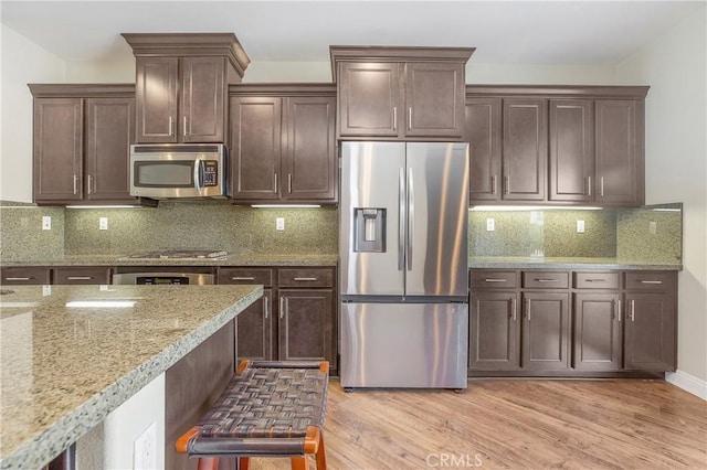 kitchen featuring light wood finished floors, decorative backsplash, appliances with stainless steel finishes, dark brown cabinets, and light stone countertops