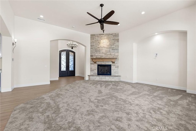 unfurnished living room with french doors, ceiling fan with notable chandelier, light hardwood / wood-style flooring, and a stone fireplace