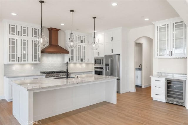 kitchen with white cabinets, wall chimney range hood, stainless steel appliances, and an island with sink