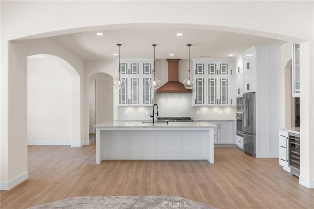 kitchen featuring pendant lighting, custom exhaust hood, a center island with sink, light hardwood / wood-style flooring, and white cabinetry