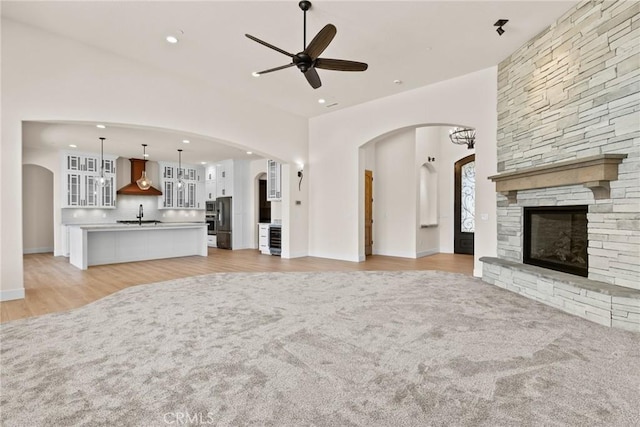 unfurnished living room with ceiling fan, sink, a stone fireplace, wine cooler, and light hardwood / wood-style floors