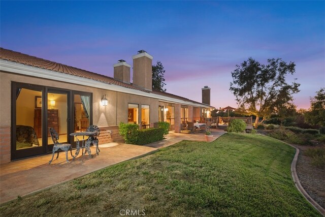 back house at dusk featuring a patio and a lawn