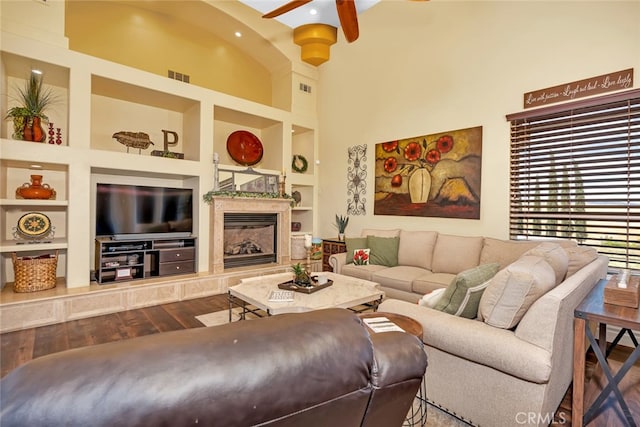 living room featuring hardwood / wood-style flooring, built in shelves, a fireplace, a towering ceiling, and ceiling fan