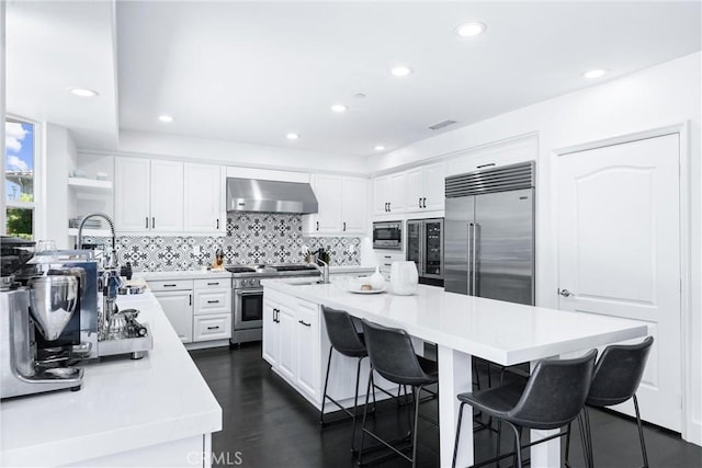 kitchen with an island with sink, a breakfast bar area, built in appliances, wall chimney range hood, and backsplash