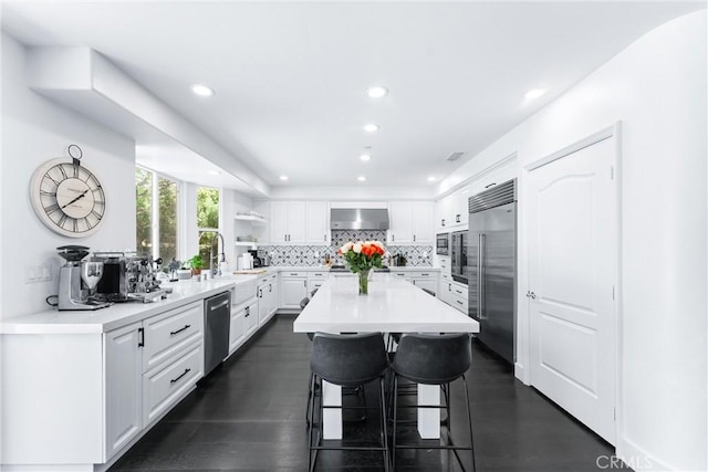 kitchen featuring white cabinetry, wall chimney range hood, appliances with stainless steel finishes, tasteful backsplash, and a kitchen bar