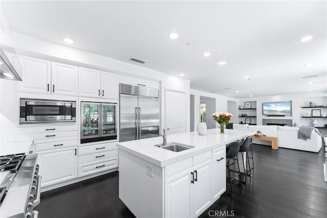 kitchen with built in appliances, a kitchen island with sink, a sink, and white cabinetry