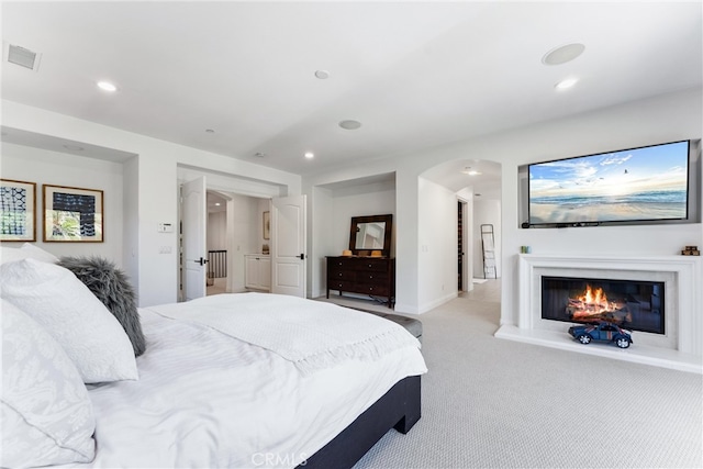 carpeted bedroom with a glass covered fireplace, visible vents, arched walkways, and recessed lighting