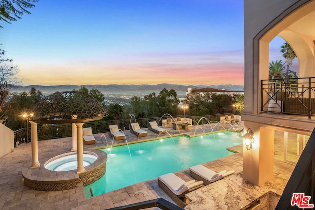 pool at dusk with an in ground hot tub, pool water feature, a mountain view, and a patio