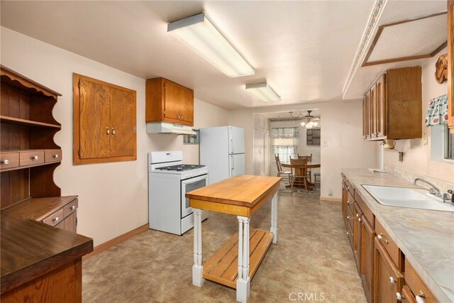 kitchen featuring brown cabinetry, a sink, white appliances, under cabinet range hood, and baseboards