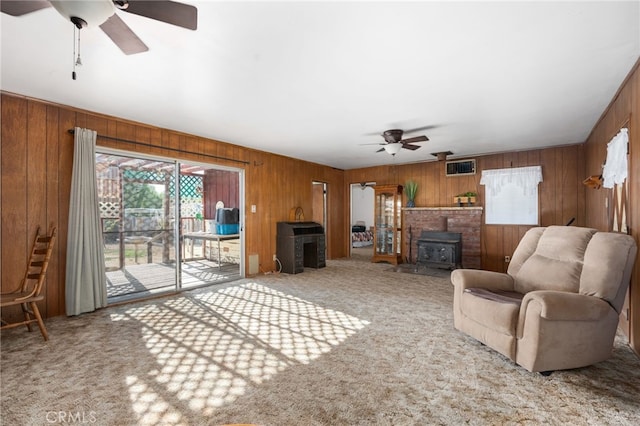 living area with wooden walls, carpet flooring, visible vents, a ceiling fan, and a wood stove