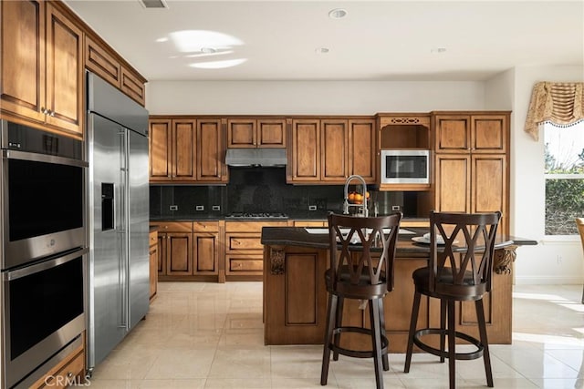 kitchen featuring light tile patterned floors, decorative backsplash, a kitchen island with sink, a kitchen breakfast bar, and built in appliances