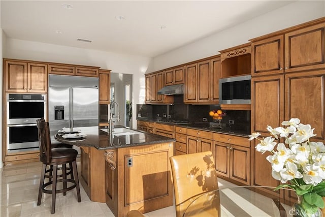 kitchen featuring decorative backsplash, a kitchen island with sink, dark stone countertops, built in appliances, and sink