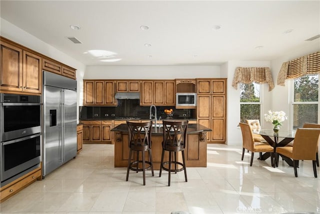 kitchen with a breakfast bar area, backsplash, a kitchen island with sink, light tile patterned flooring, and built in appliances