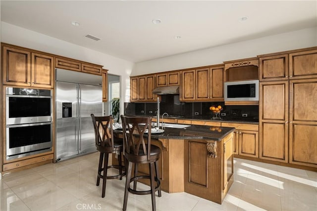 kitchen featuring light tile patterned floors, an island with sink, built in appliances, and dark stone countertops