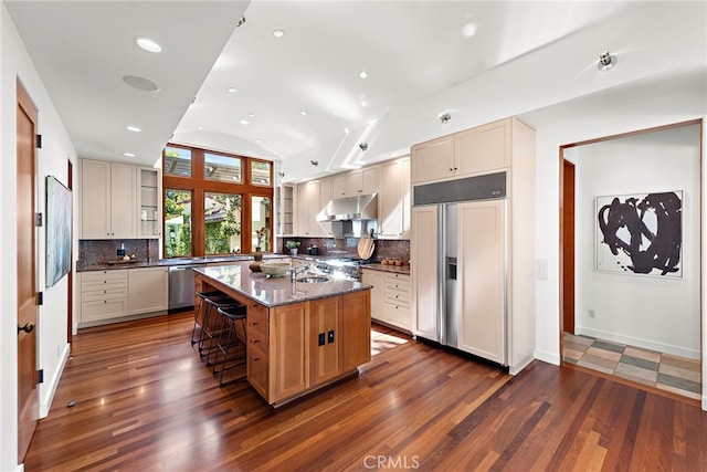 kitchen featuring dark stone counters, dark wood finished floors, dishwasher, under cabinet range hood, and paneled fridge