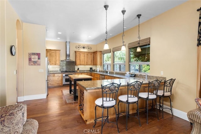 kitchen with a peninsula, dark wood-style flooring, wall chimney range hood, appliances with stainless steel finishes, and backsplash