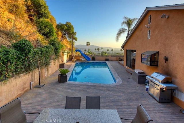 pool at dusk featuring grilling area, a mountain view, a patio area, and a water slide