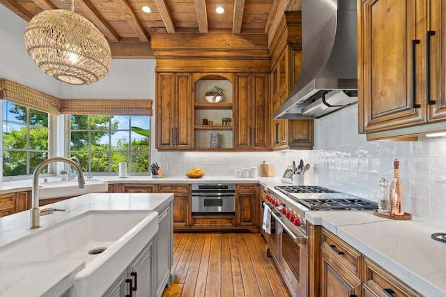 kitchen with wall chimney range hood, beam ceiling, sink, appliances with stainless steel finishes, and light stone counters